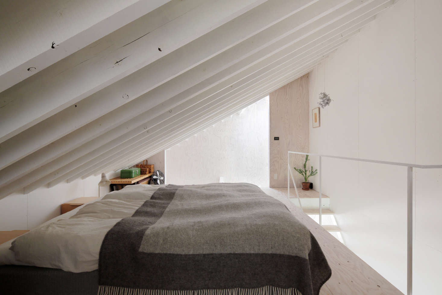 The bed is angled under the eaves next to a sloping side window. Building codes in Japan allow for entirely open stairs, such as this simple steel railing.
