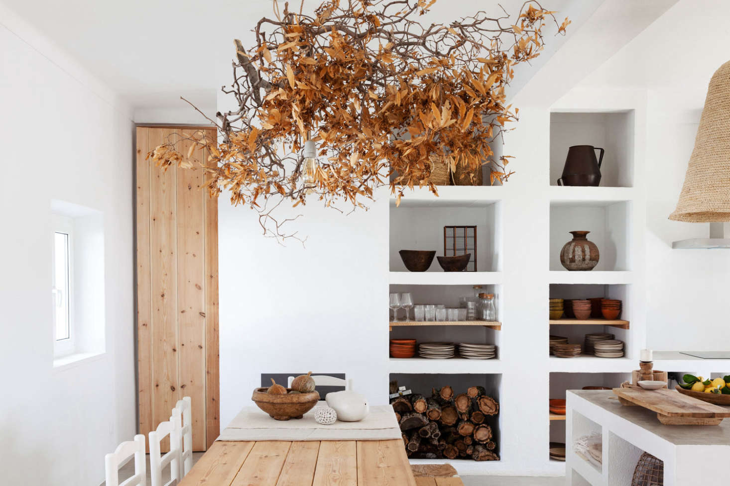 The guest house kitchen, with sculptural white shelves, a wood-plank table, and a tangle of dried leaves hung above. (For a similar autumnal look, see Bringing in the Sheaves: Harvest Table Inspiration from Caro of Somerset.)
