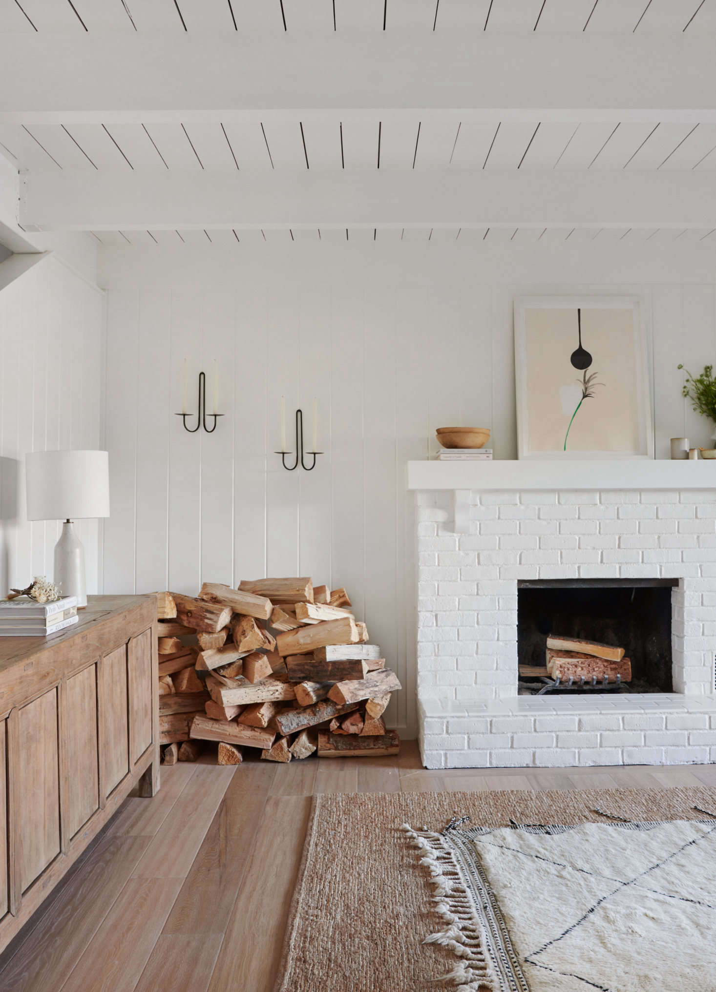 The original fireplace, painted white, is paired with layered rugs and a generous stack of firewood. The wall-mounted candle holders are from June Home Supply. (They