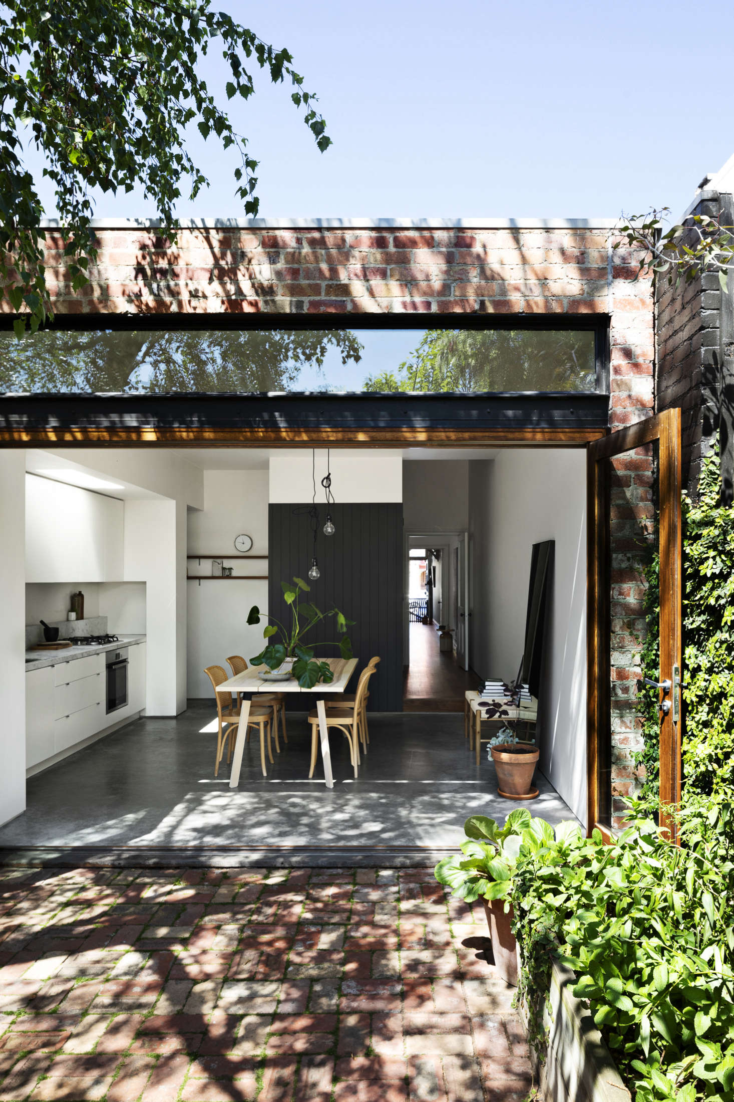 The center of the house is a bright kitchen and dining area with custom bifold doors and a transom window that fully connects indoors to out. The shotgun hallway was created by flipping the position of the original hall. The charcoal paneled partition hides the fridge and laundry.