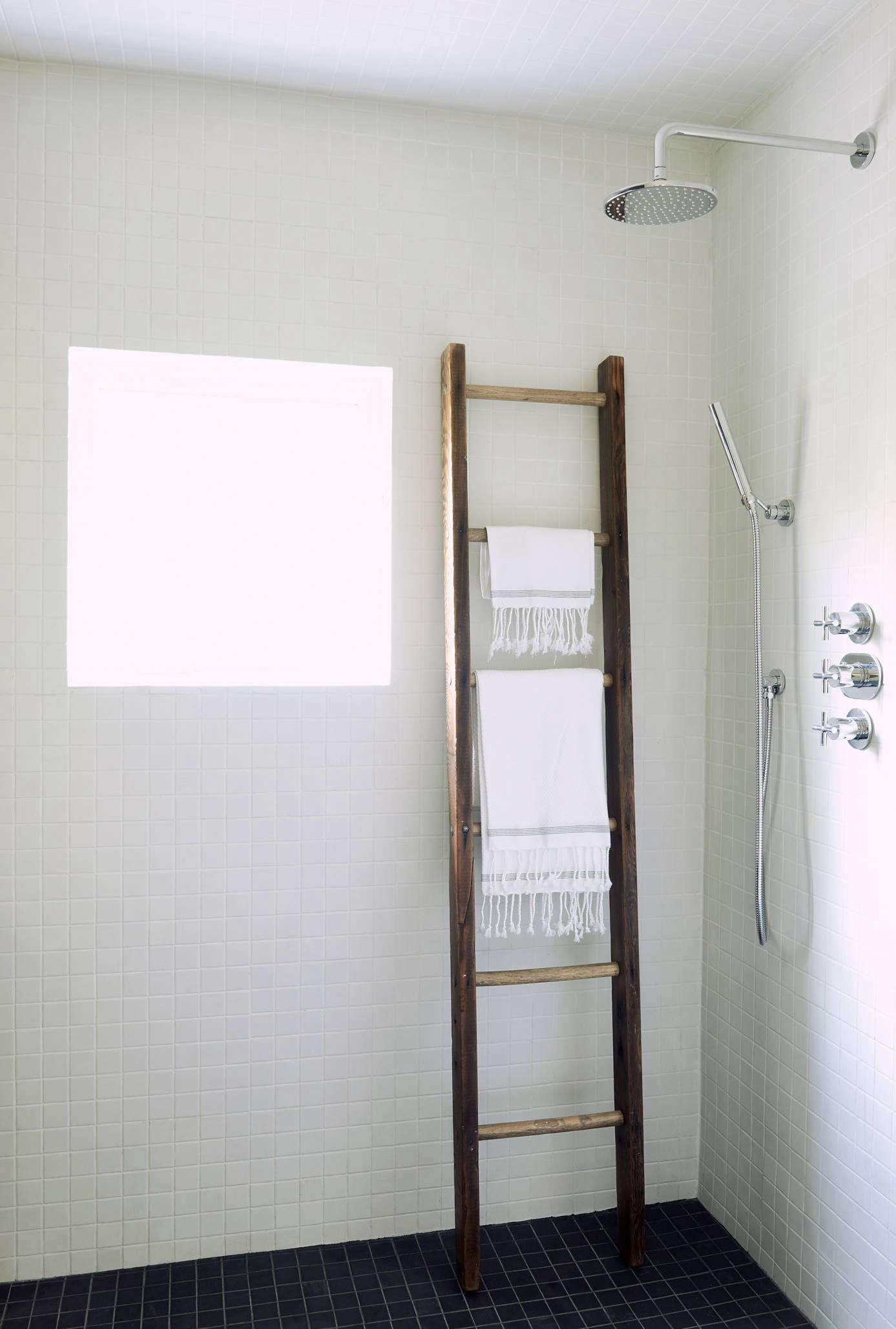 White mosaic tiles cover the rest of the walls. A vintage wooden ladder, used as a towel holder, adds some warmth to the room. The shower fixtures are by Signature Hardware.