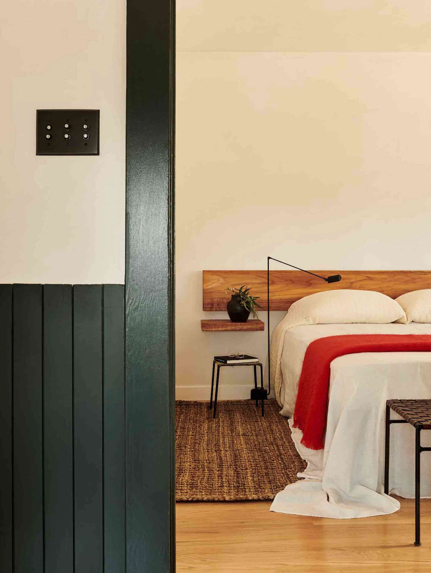 The main bedroom features a simple headboard and matching floating shelves, a red mohair blanket from Mantas Ezcaray, and custom cotton bedding from Oaxaca. (Note the dramatic floor-length pillow cases.) Outside the room is an oil-rubbed bronze switch plate from Rejuvenation.