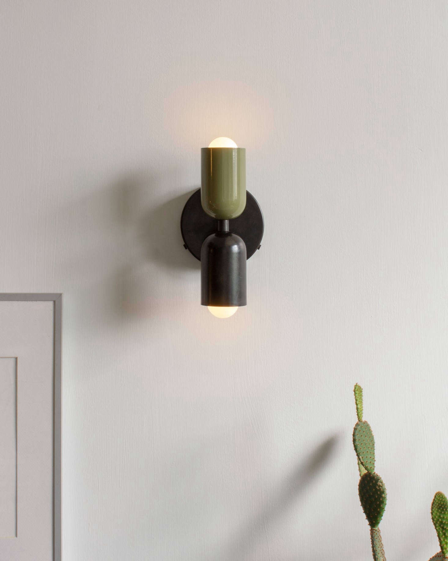 The Up Down Sconce with a Reed Green top shade and Black bottom shade. The light is 4 7/8 in wide by 9 5/8 in tall and is available hardwired or with a plug-in cord.